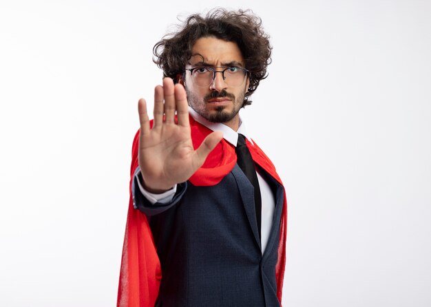 Homem jovem e confiante super-herói caucasiano com óculos ópticos, usando um terno com capa vermelha, gesticulando para parar