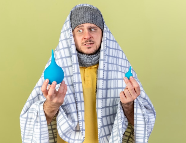 Foto grátis homem jovem e confuso, loiro, doente, eslavo, usando chapéu de inverno e lenço embrulhado em manta, olha para enemas isolados na parede verde oliva com espaço de cópia