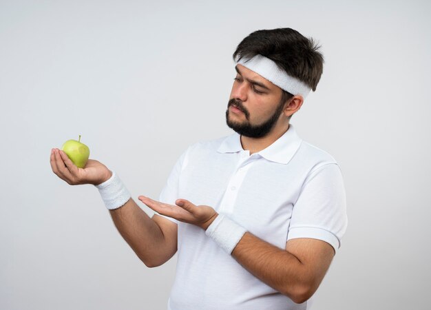 Homem jovem e desportivo descontente com fita na cabeça e pulseira segurando e aponta com a mão na maçã isolada na parede branca
