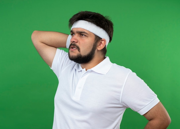 Homem jovem e esportivo preocupado, olhando para o lado usando bandana e pulseira, colocando a mão no pescoço isolado em verde