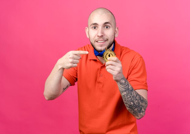 Homem jovem e esportivo satisfeito usando e aponta para medalha isolada na parede rosa