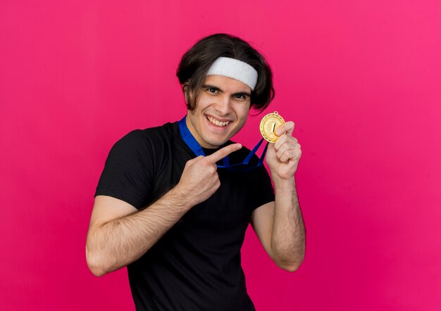 Homem jovem e esportivo satisfeito usando roupas esportivas e bandana com medalha de ouro no pescoço apontando com o dedo indicador para a medalha feliz sorrindo