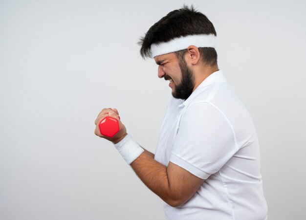 Homem jovem e esportivo tenso em vista de perfil usando bandana e pulseira, fazendo exercícios com halteres isolados na parede branca com espaço de cópia