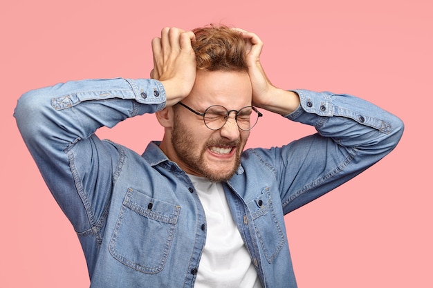 Homem jovem e estressante com a barba por fazer sofre de dor de cabeça, mantém as mãos nas têmporas, cerra os dentes, tem uma dor terrível, fecha os olhos