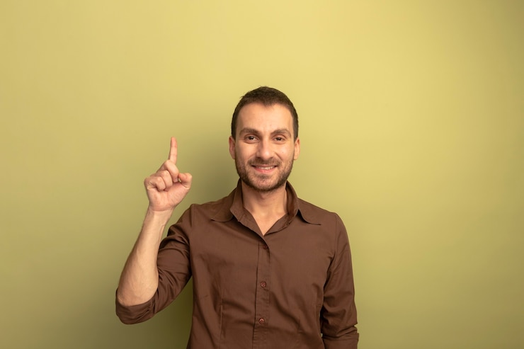 Foto grátis homem jovem e sorridente, caucasiano, olhando para a câmera, apontando para cima, isolado em um fundo verde oliva com espaço de cópia