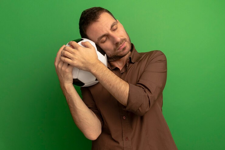 Foto grátis homem jovem e tranquilo, caucasiano, segurando uma bola de futebol, colocando a cabeça nela com os olhos fechados, isolada em um fundo verde com espaço de cópia