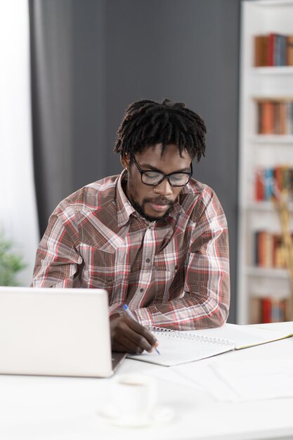 Homem jovem estudante africano fazendo anotações trabalhando em casa trabalhando no laptop ou ouvindo professor ou chefe durante a videochamada