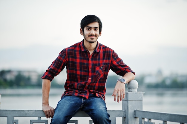 Foto grátis homem jovem estudante indiano na camisa quadriculada e jeans sentado no corrimão contra o lago