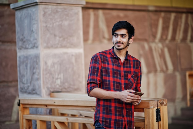 Foto grátis homem jovem estudante indiano na camisa xadrez vermelha e jeans posou na cidade com o celular nas mãos