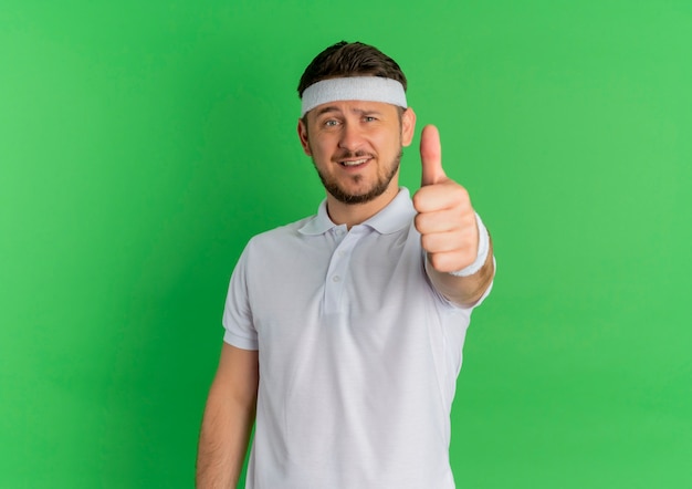 Homem jovem fitness com camisa branca e bandana sorrindo, olhando para a frente, mostrando os polegares em pé sobre a parede verde
