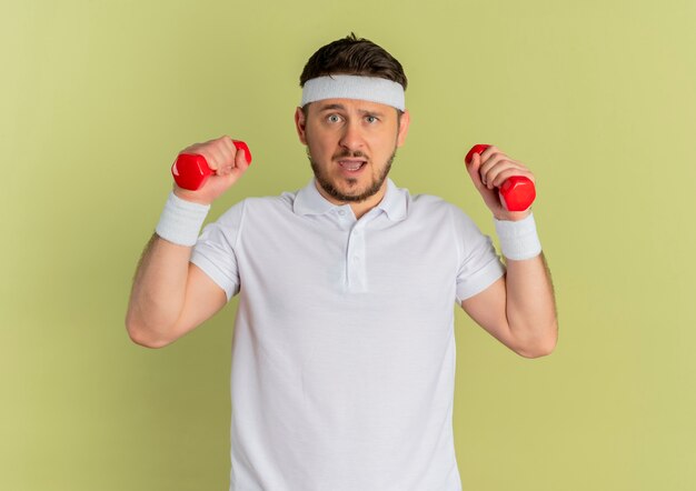 Homem jovem fitness com camisa branca e faixa na cabeça, malhando com halteres, parecendo confuso em pé sobre um muro de oliveiras