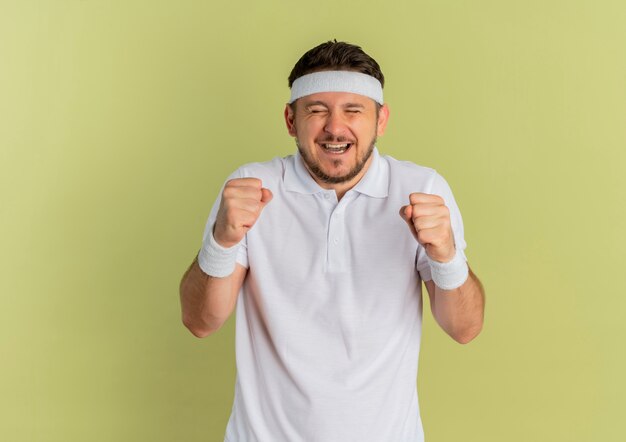 Homem jovem fitness com camisa branca e faixa na cabeça, punhos cerrados, feliz e animado, regozijando-se com seu sucesso em pé sobre fundo verde-oliva