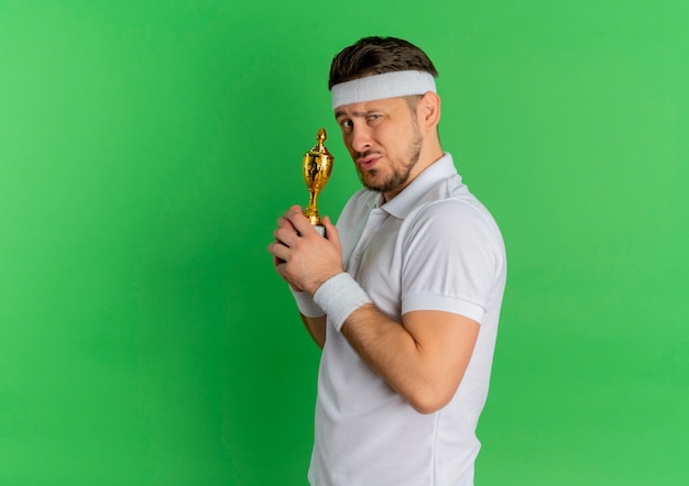 Homem jovem fitness com camisa branca e fita na cabeça segurando um troféu, parecendo confiante em pé sobre fundo verde