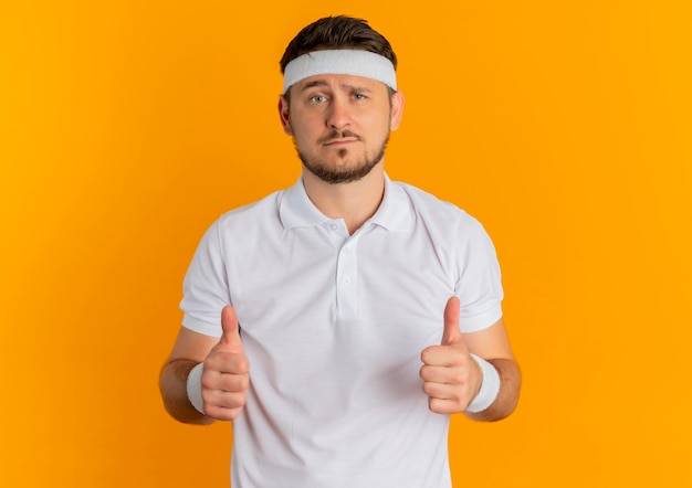 Homem jovem fitness com camisa branca e tiara mostrando os polegares para cima, olhando para a frente em pé sobre a parede laranja