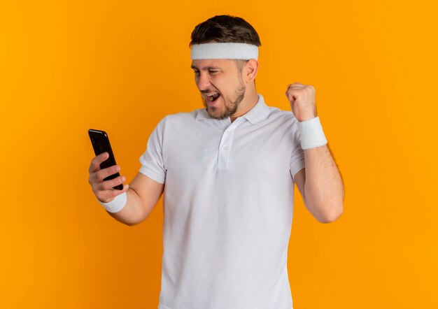 Homem jovem fitness em camisa branca com tiara golding smartphone punho cerrado feliz e animado em pé sobre fundo laranja