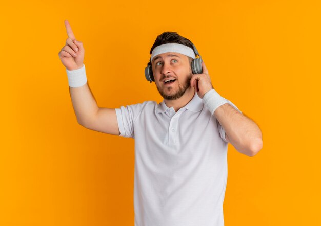 Homem jovem fitness em uma camisa branca com bandana e fones de ouvido, parecendo surpreso e feliz, mostrando o dedo indicador tendo uma ótima ideia em pé sobre a parede laranja