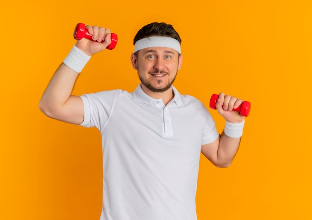 Homem jovem fitness em uma camisa branca com bandana fazendo exercícios com halteres olhando para a câmera com um sorriso no rosto em pé sobre um fundo laranja