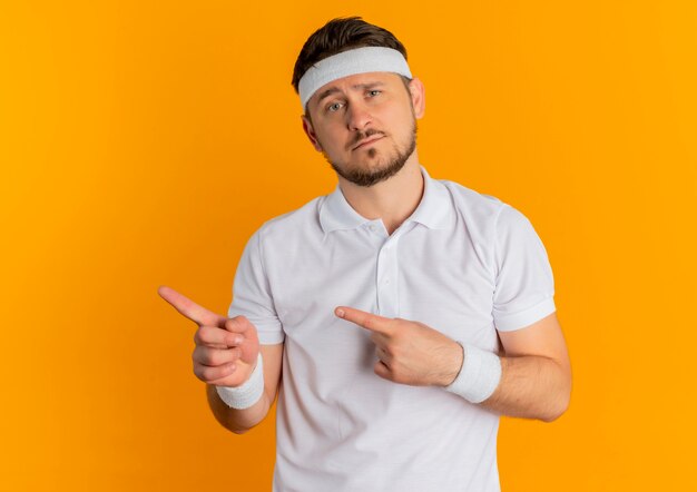 Homem jovem fitness em uma camisa branca com bandana olhando para a frente com uma expressão triste no rosto apontando com os dedos para o lado em pé sobre a parede laranja