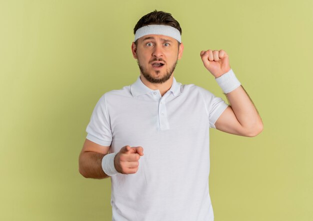 Homem jovem fitness em uma camisa branca com uma faixa na cabeça apontando com o dedo para a frente levantando o punho parecendo surpreso em pé sobre um muro de oliveiras