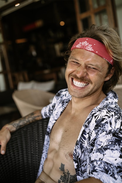 Homem jovem hippie bonito em uma bandana vermelha, com um torso nu em um café em bali. homem feliz sorrindo