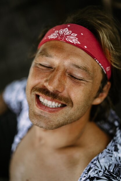Homem jovem hippie bonito em uma bandana vermelha, com um torso nu em um café em Bali. Homem feliz sorrindo