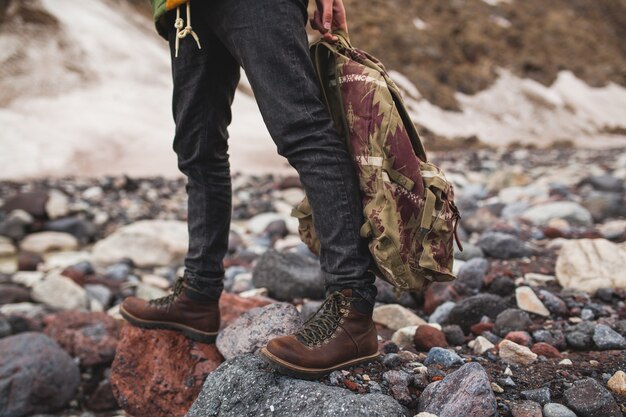 Homem jovem hippie, caminhando à beira do rio, natureza selvagem, férias de inverno, segurando uma mochila nas mãos, detalhes de close up