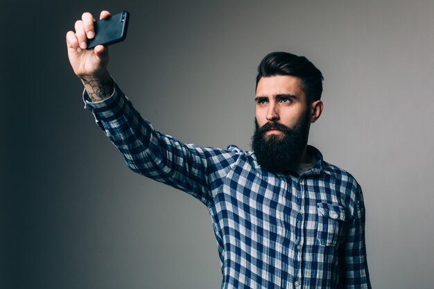 Homem jovem hippie com barba comprida tomando selfie com as mãos na barba em pé na parede cinza