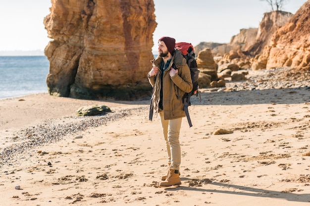 Homem jovem hippie viajando com mochila na costa do mar de outono, vestindo jaqueta e chapéu.