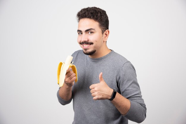 Homem jovem positivo segurando a banana e aparecendo um polegar.