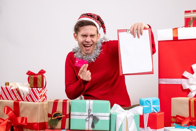 Homem jovem segurando um cartão e uma prancheta sentado em frente aos presentes de Natal