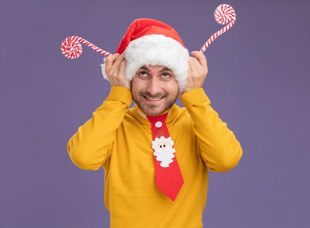 Homem jovem sorridente, caucasiano, usando gravata e chapéu de Natal, segurando bengalas doces de Natal, olhando para cima, fazendo chifres de rena com bengalas isoladas no fundo roxo