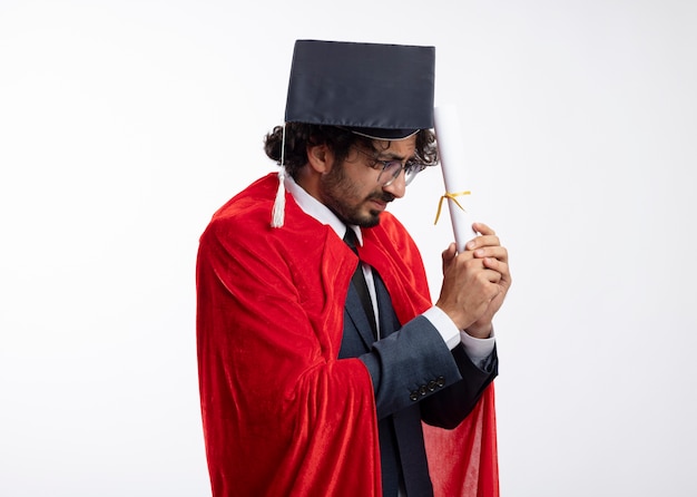 Foto grátis homem jovem super-herói caucasiano descontente com óculos ópticos, vestindo um terno com capa vermelha e boné de formatura segurando e olhando para diploma