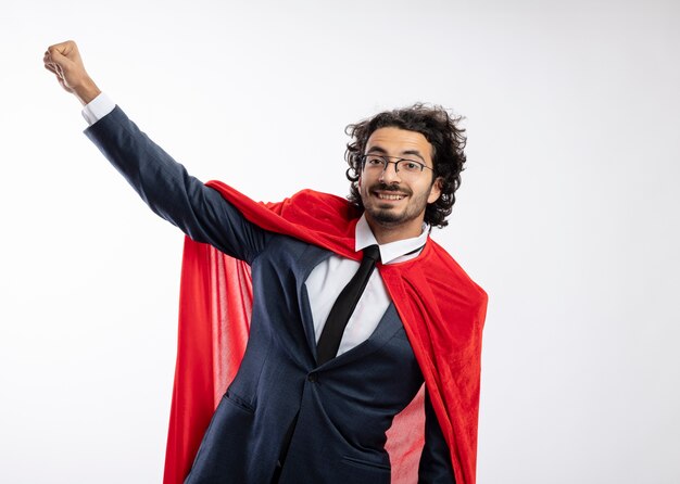 Homem jovem super-herói caucasiano sorridente usando óculos ópticos e terno com capa vermelha levanta o punho isolado no fundo branco com espaço de cópia