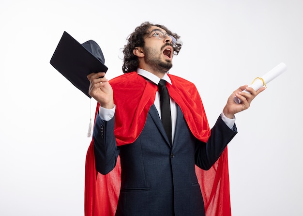 Homem jovem super-herói chocado usando óculos óticos, usando terno com capa vermelha, segurando um boné de formatura e diploma isolado na parede branca