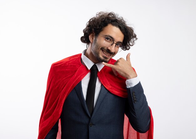 Homem jovem super-herói sorridente usando óculos ópticos e terno com manto vermelho. Gestos me chamam de sinal isolado na parede branca