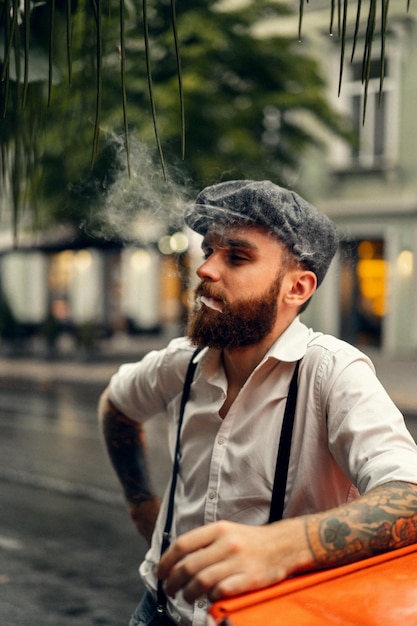 Homem jovem tatuado barbudo em um café na rua fuma um cigarro. cara romântico em um boné de camisa branca e suspensórios na cidade. peaky blinders. retrô na moda antiga.