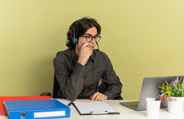 Homem jovem trabalhador de escritório confiante com fones de ouvido em óculos ópticos, sentado na mesa com ferramentas de escritório, usando e olhando para o laptop coloca a mão no queixo isolado no fundo verde com espaço de cópia
