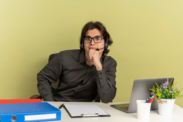 Homem jovem trabalhador de escritório confiante com fones de ouvido em óculos ópticos, sentado na mesa com ferramentas de escritório usando laptop coloca a mão no queixo olhando para a câmera isolada em um fundo verde com espaço de cópia