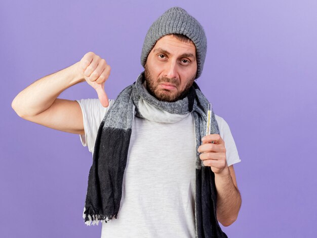 Homem jovem triste e doente com chapéu de inverno com lenço segurando o termômetro e mostrando o polegar isolado no roxo