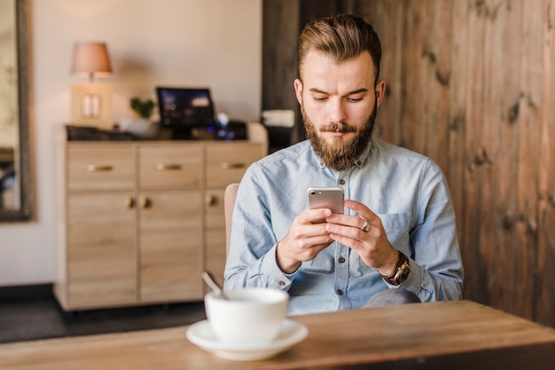 Homem jovem, usando, telefone móvel, com, xícara café, escrivaninha