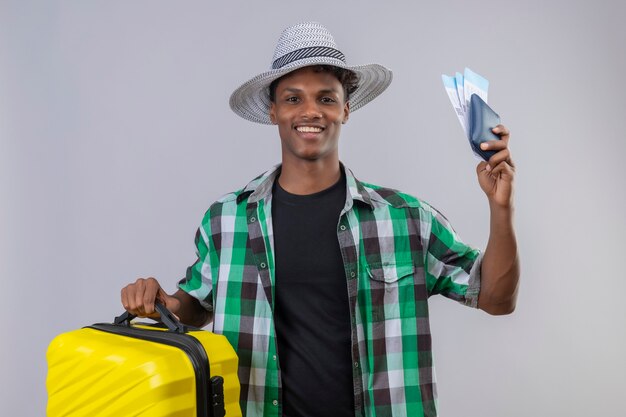 Homem jovem viajante afro-americano com chapéu de verão e mala segurando passagens aéreas, sorrindo alegremente, positivo e feliz.