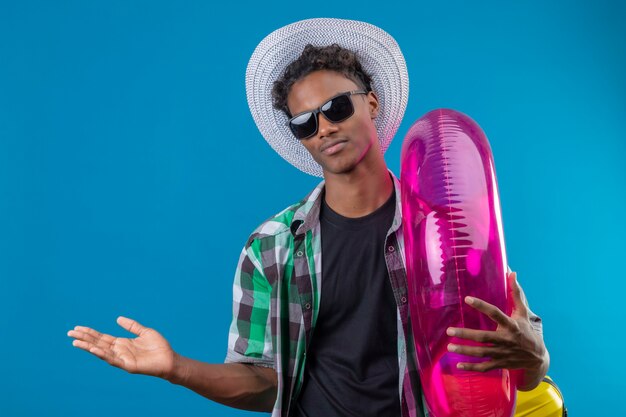 Homem jovem viajante afro-americano com chapéu de verão, usando óculos escuros pretos, segurando um anel inflável, apresentando com o braço da mão, copie o espaço olhando para a câmera com um sorriso confiante.