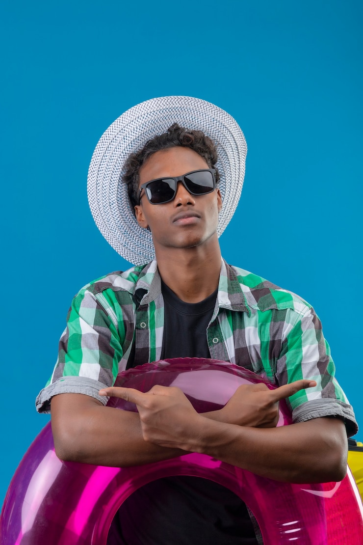 Foto grátis homem jovem viajante afro-americano com chapéu de verão usando óculos escuros pretos segurando um anel inflável com as mãos cruzadas apontando para os lados com os dedos