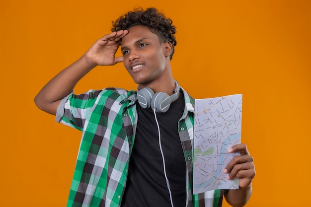 Homem jovem viajante afro-americano com fones de ouvido segurando um mapa, olhando de lado perplexo em pé sobre um fundo laranja
