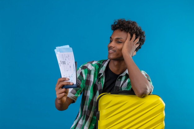 Homem jovem viajante afro-americano com mala segurando passagens aéreas, olhando para eles com um sorriso confiante no rosto, satisfeito