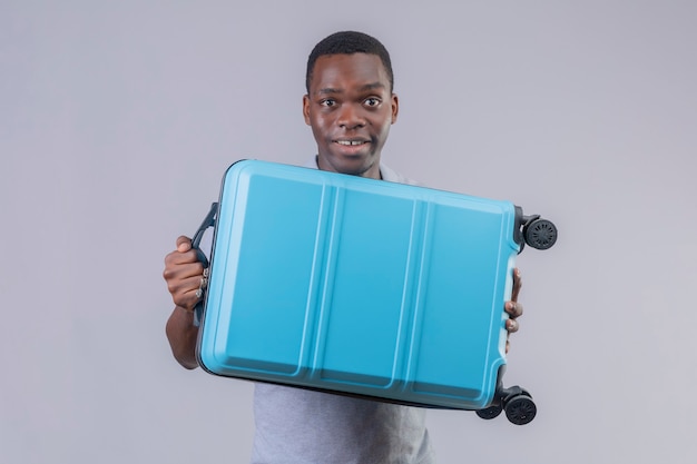 Homem jovem viajante afro-americano com uma camisa pólo cinza segurando uma mala azul parecendo surpreso e feliz