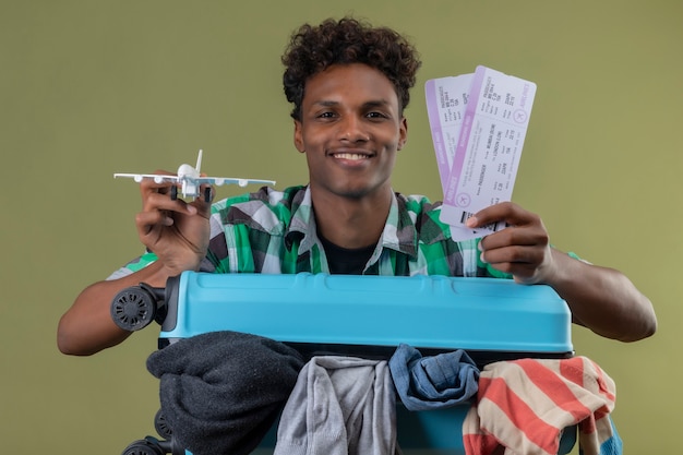 Homem jovem viajante afro-americano em pé com uma mala cheia de roupas segurando passagens aéreas e um avião de brinquedo olhando para a câmera sorrindo feliz e positivo sobre o fundo verde