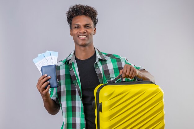 Homem jovem viajante afro-americano em pé com uma mala segurando passagens aéreas, sorrindo alegremente positivo e feliz sobre fundo branco.