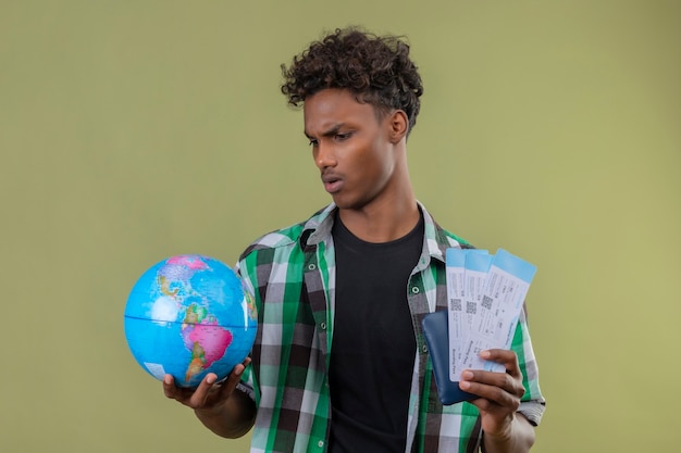 Homem jovem viajante afro-americano segurando passagens aéreas e um globo olhando para ele com uma expressão séria no rosto franzindo a testa em pé sobre um fundo verde