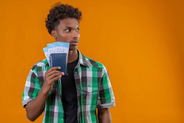 Homem jovem viajante afro-americano segurando passagens aéreas, olhando para o lado com expressão de medo no rosto em pé sobre um fundo laranja